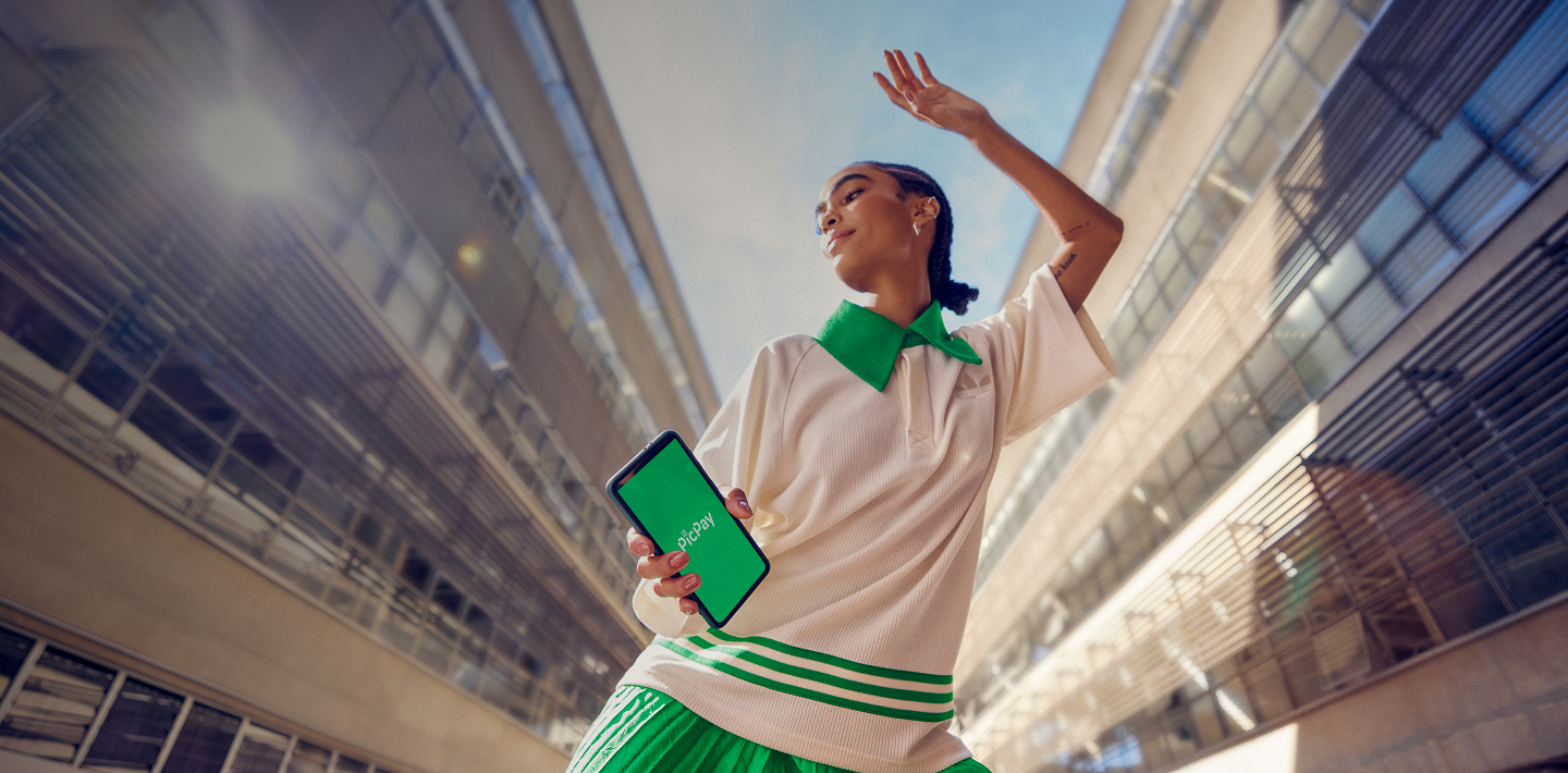 Mulher em pose de dança descontraída, usando uma camisa branca com listras verdes, segurando um celular que tem a logo do PicPay em um fundo verde.