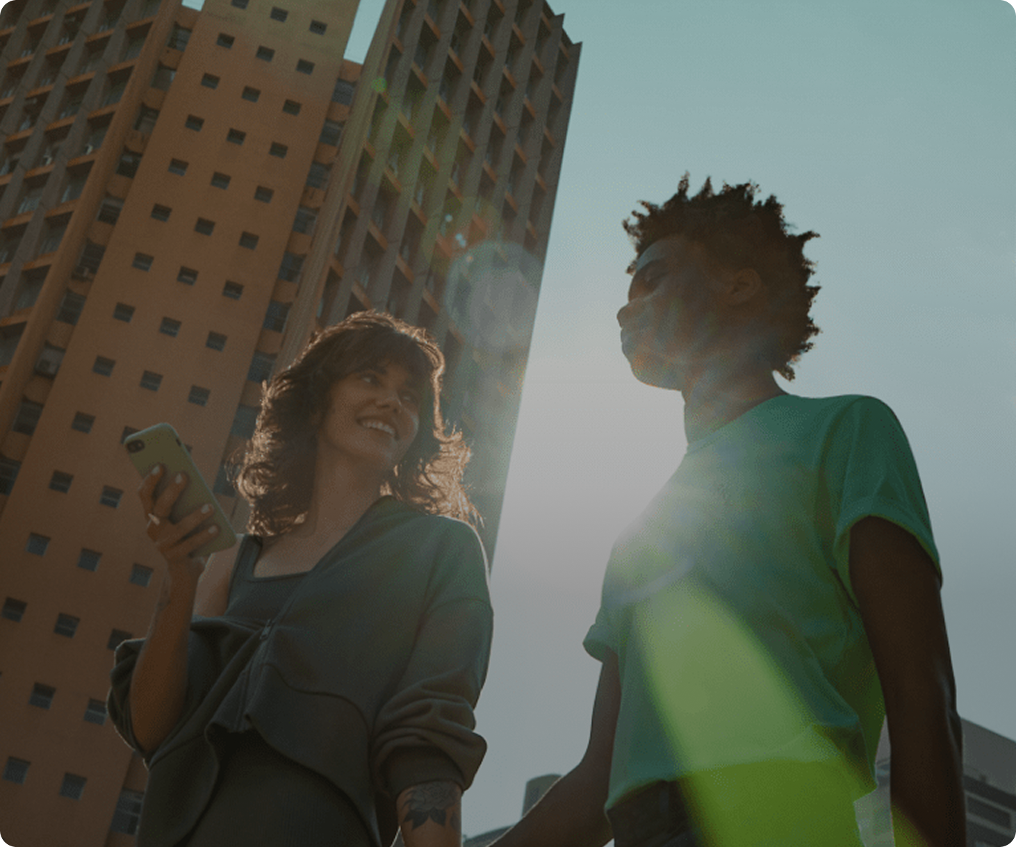 Um homem e uma mulher olhando um para outro sorrindo, a mulher com o celular na mão e um prédio grande atras dela