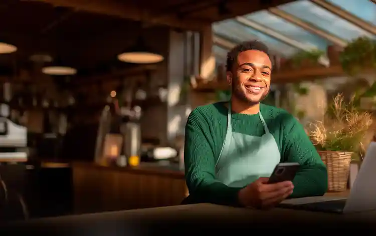 Homem usando avental em ambiente de café, sorrindo enquanto utiliza o celular ao lado de um notebook, representando empreendedor usando soluções digitais para o negócio.