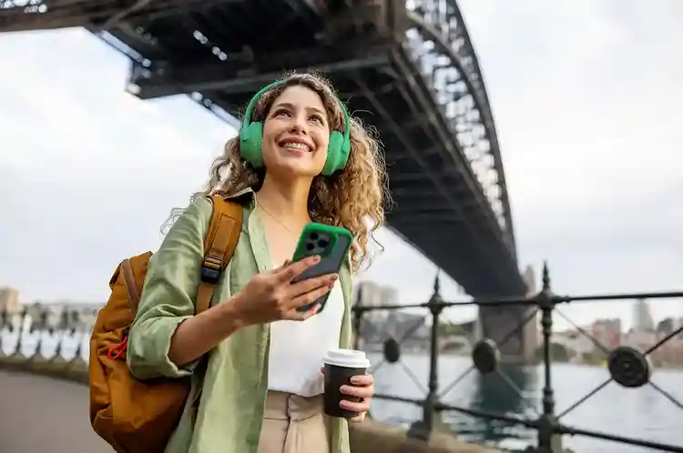 Mulher sorrindo enquanto caminha, usando fones de ouvido verdes e segurando um celular e um copo de café, com uma ponte e um rio ao fundo.