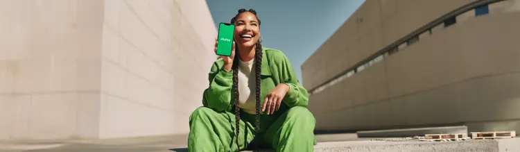 Pessoa sentada em área externa de concreto, usando roupas verdes e segurando um smartphone com o logo do PicPay.