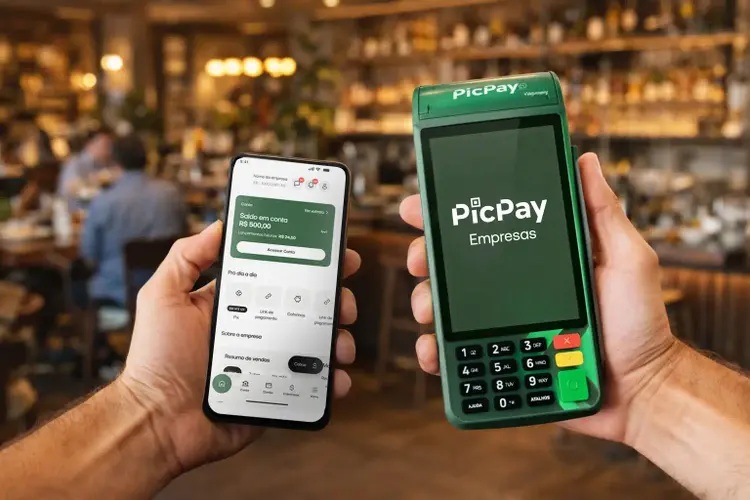 Uma mão segura um smartphone com o app do Picpay e a outra segura uma máquina de cartão, em primeiro plano, com fundo desfocado.