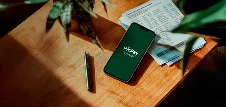 Smartphone sobre mesa de madeira exibindo o logo do PicPay Empresas, ao lado de uma caneta e documentos.