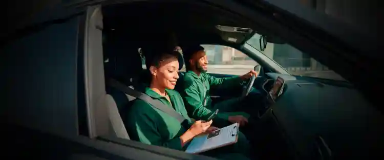 Duas pessoas sentadas nos bancos dianteiros de um carro, usando cintos de segurança, uma segura um smartphone, uma prancheta e com o cartão PicPay, enquanto a outra está ao volante.